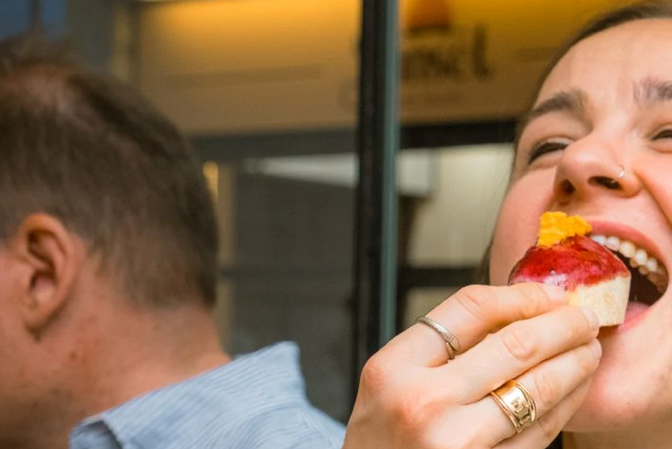 A woman happily eating a piece of food while seated indoors next to another person.