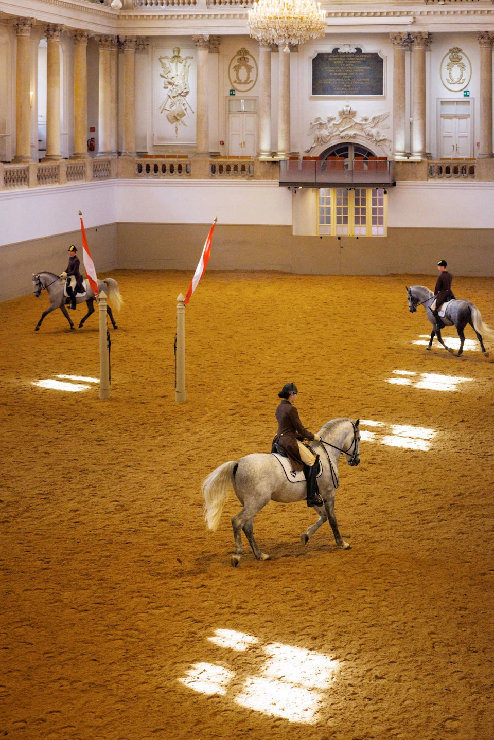 Three riders on white horses practice in an indoor arena with two red-and-white flags on poles.