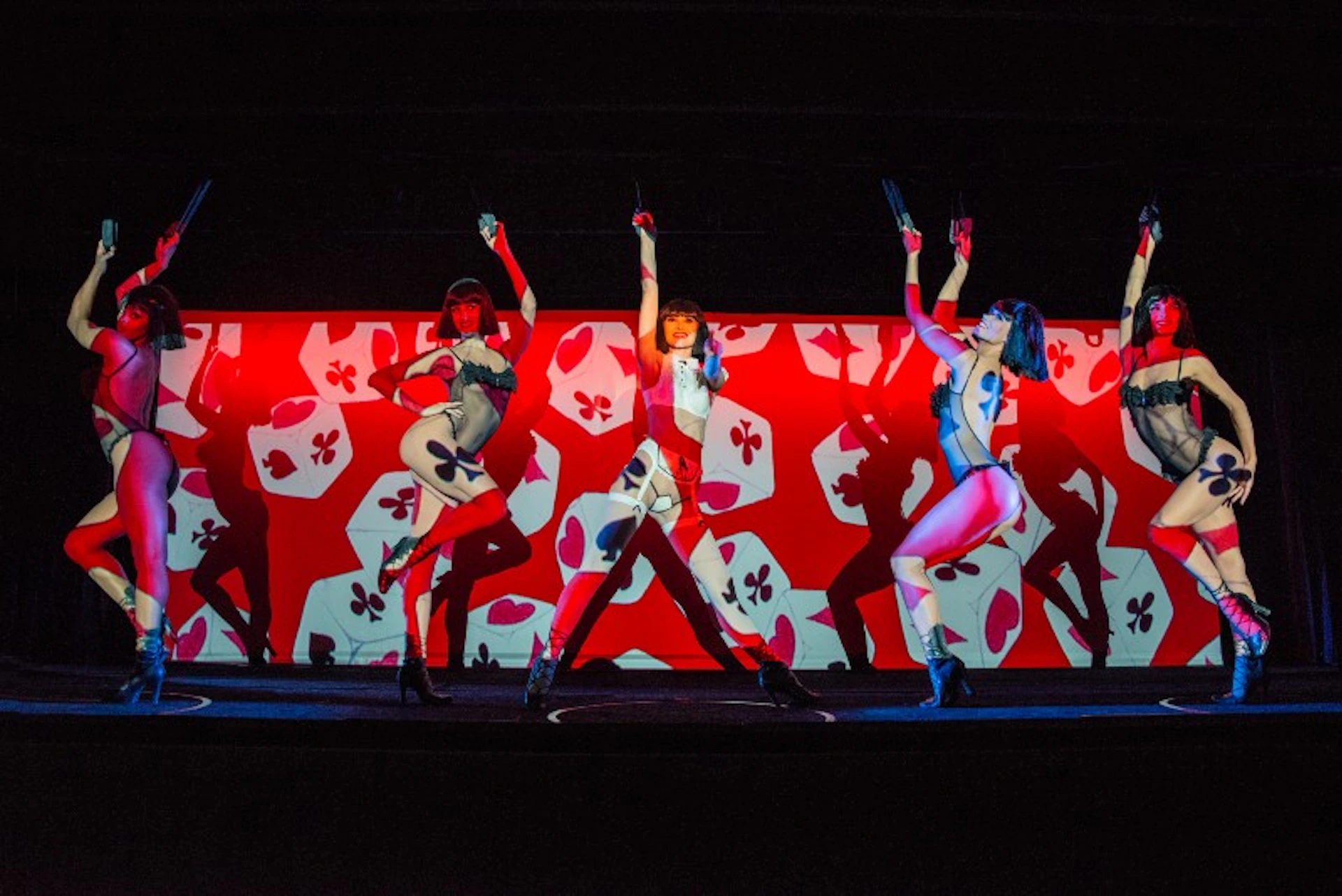 Three dancers in elaborate outfits perform on a stage with a red and white floral patterned backdrop.