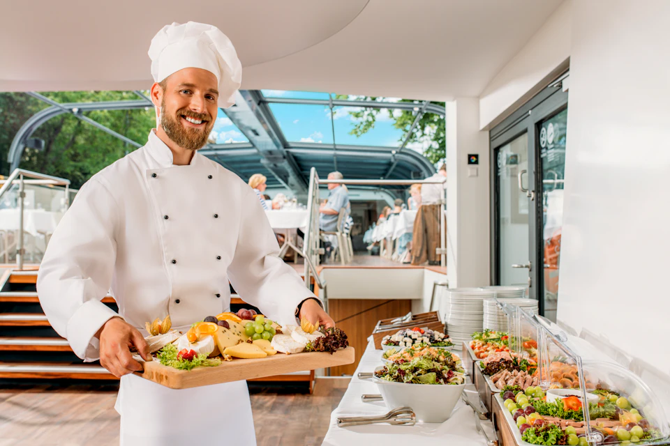 A chef in a white uniform and hat holds a wooden tray with assorted food. Indoor dining area with people in the background.