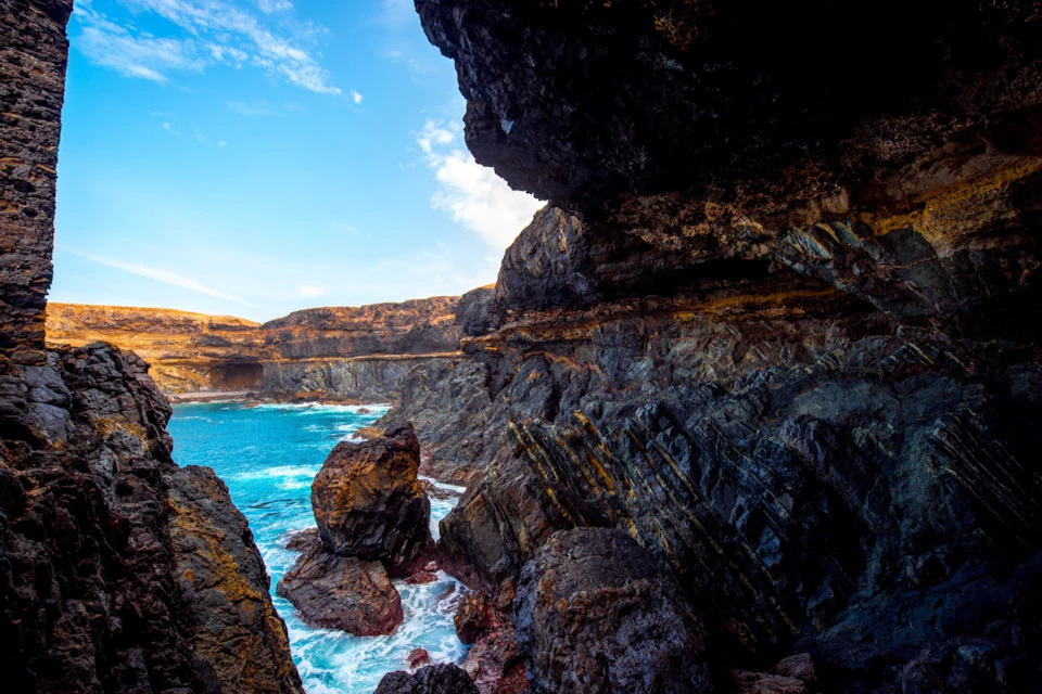 Rugged coastal cliffs with dark volcanic rock formations and turquoise sea under a partly cloudy blue sky.