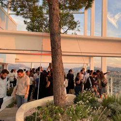 People mingling at an outdoor gathering with a large tree, modern architecture, and scenic city view in the background.