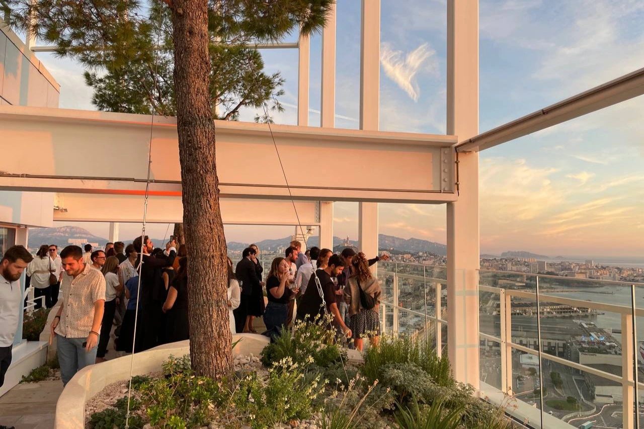 People mingling at an outdoor gathering with a large tree, modern architecture, and scenic city view in the background.