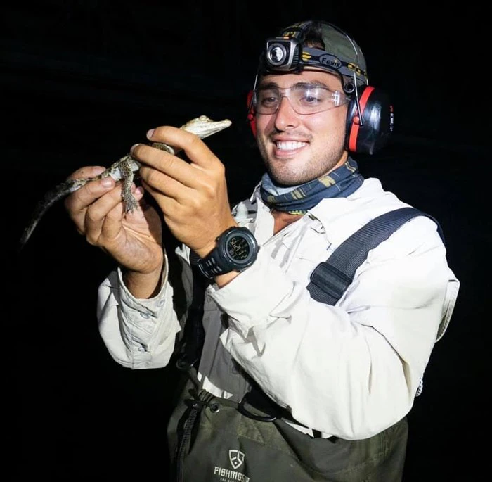 A person wearing outdoor gear and a headlamp smiles while holding a small reptile at night.