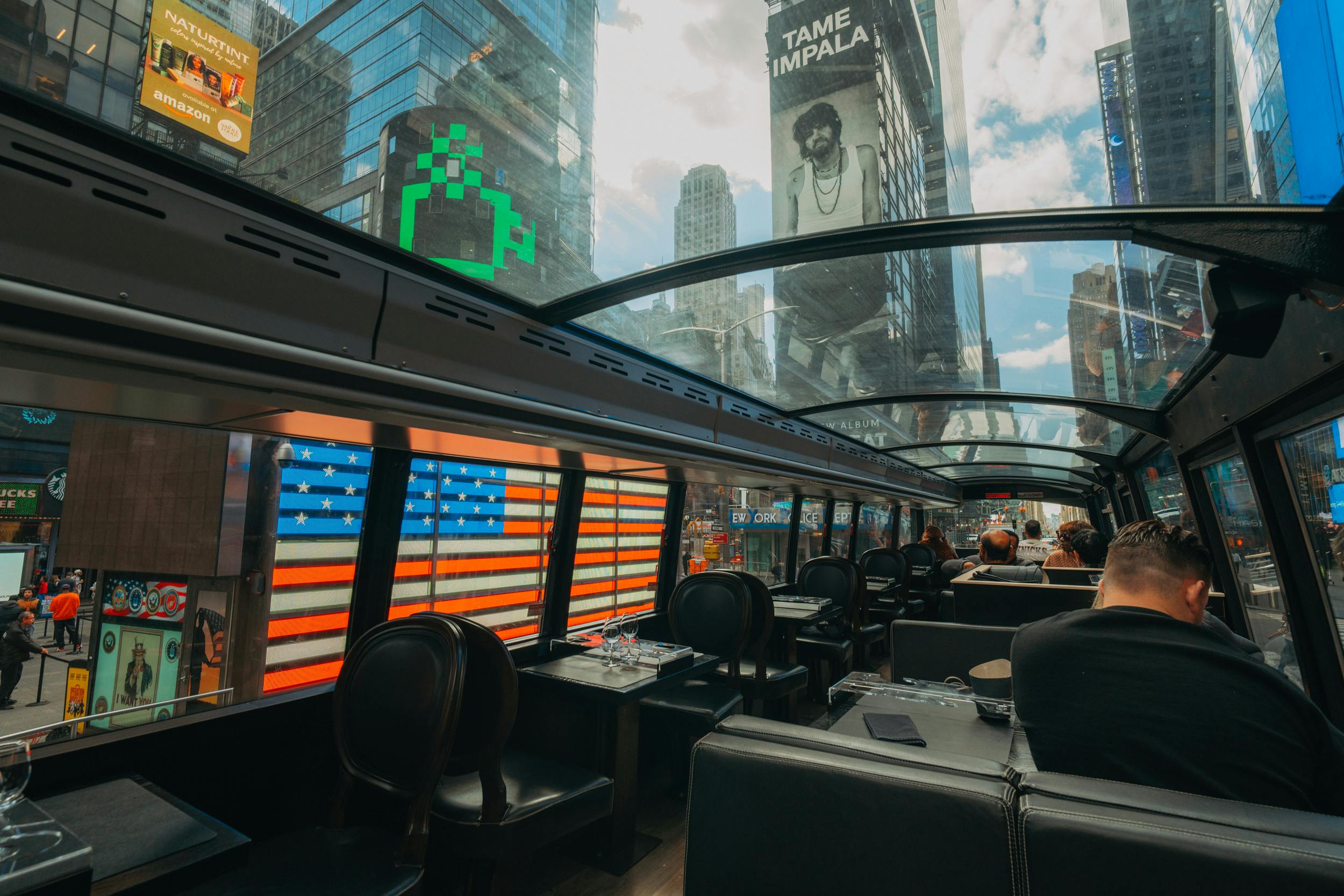 In een bus met een glazen dak, met uitzicht op gebouwen in de stad, een reclamebord en een patroon van Amerikaanse vlaggen op de ramen van de bus.