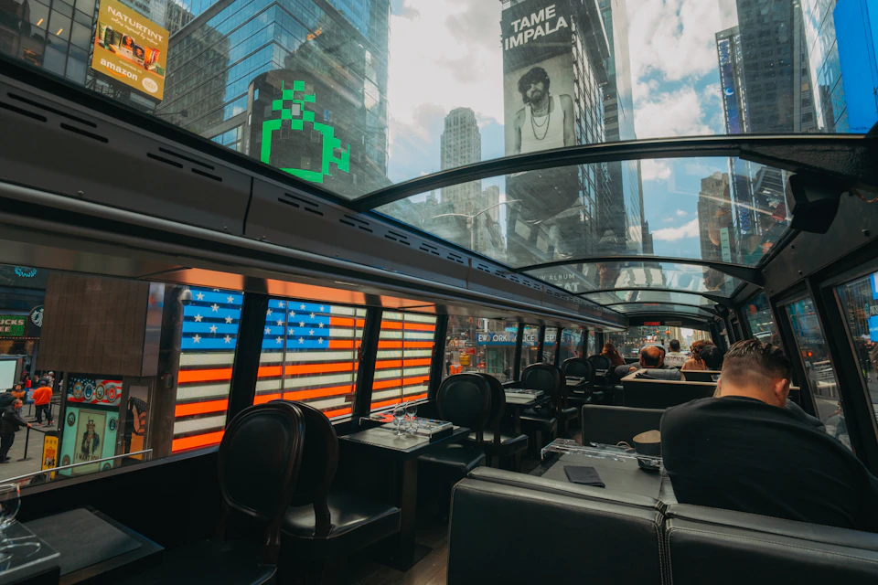 Inside a bus with a glass roof, showing a view of city buildings, a billboard, and an American flag pattern on the bus windows.