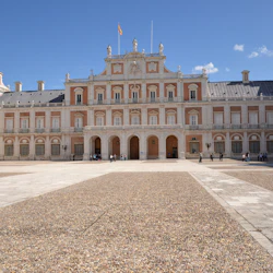 Stort palats med utsmyckad fasad, välvda entréer och två välvda torn. Öppen innergård med spridda besökare under en klar himmel.