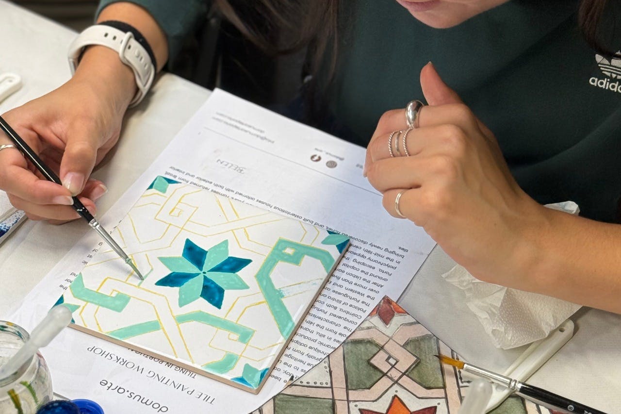 Eine Person, die ein geometrisches Muster auf eine Fliese malt, wobei ein Papier und andere Werkzeuge auf dem Tisch liegen.
