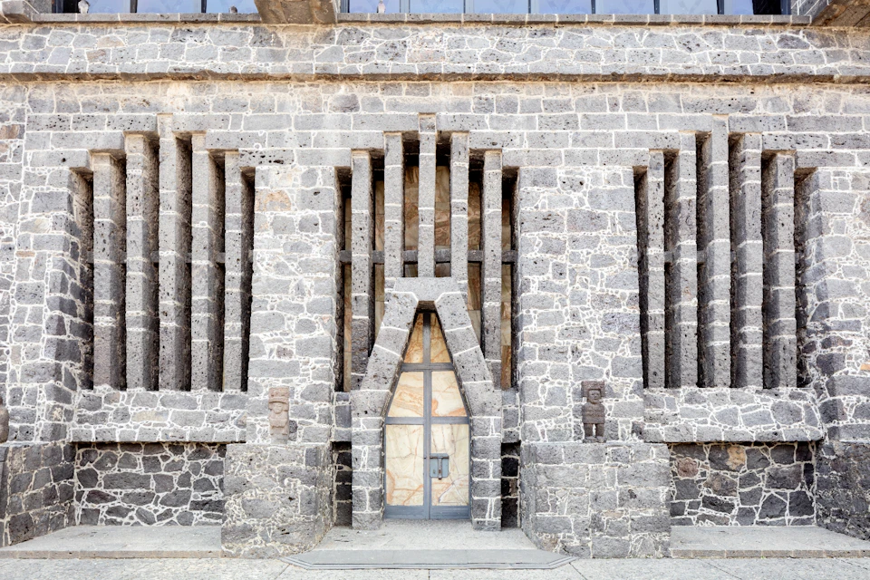 Gray stone facade with a geometric pattern, narrow vertical windows, and a central door framed by angular stonework.