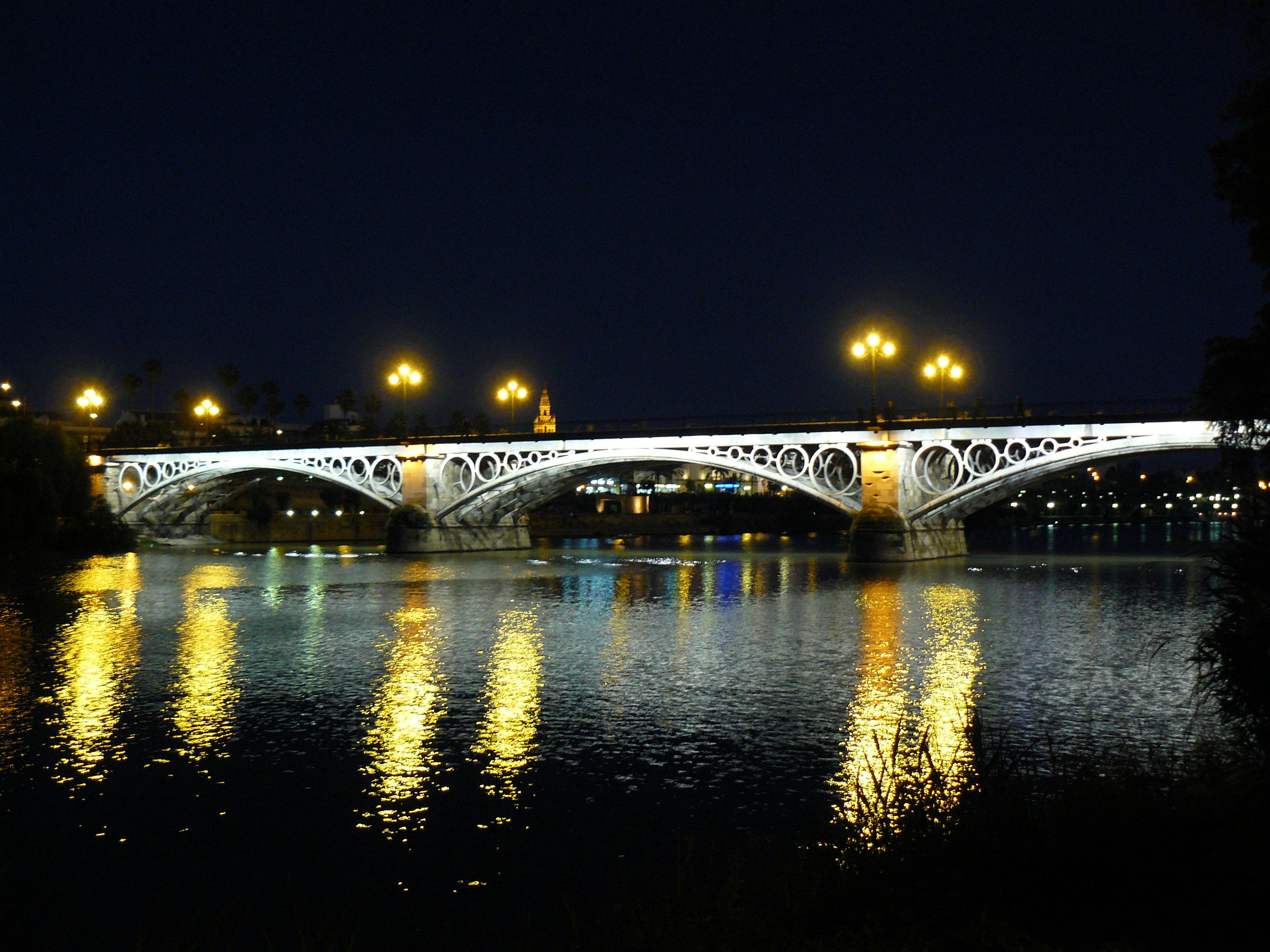 Освещенный арочный мост ночью с огнями, отражающимися от воды внизу, видимый городской пейзаж на заднем плане.
