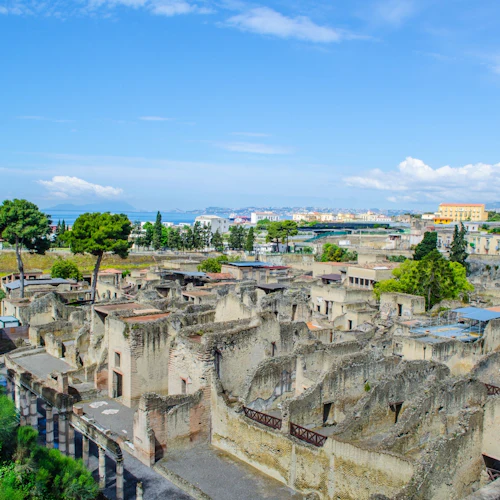 Forntida ruiner med stenstrukturer och pelare, omgivna av grönska, med en stad och hav i bakgrunden under en blå himmel.