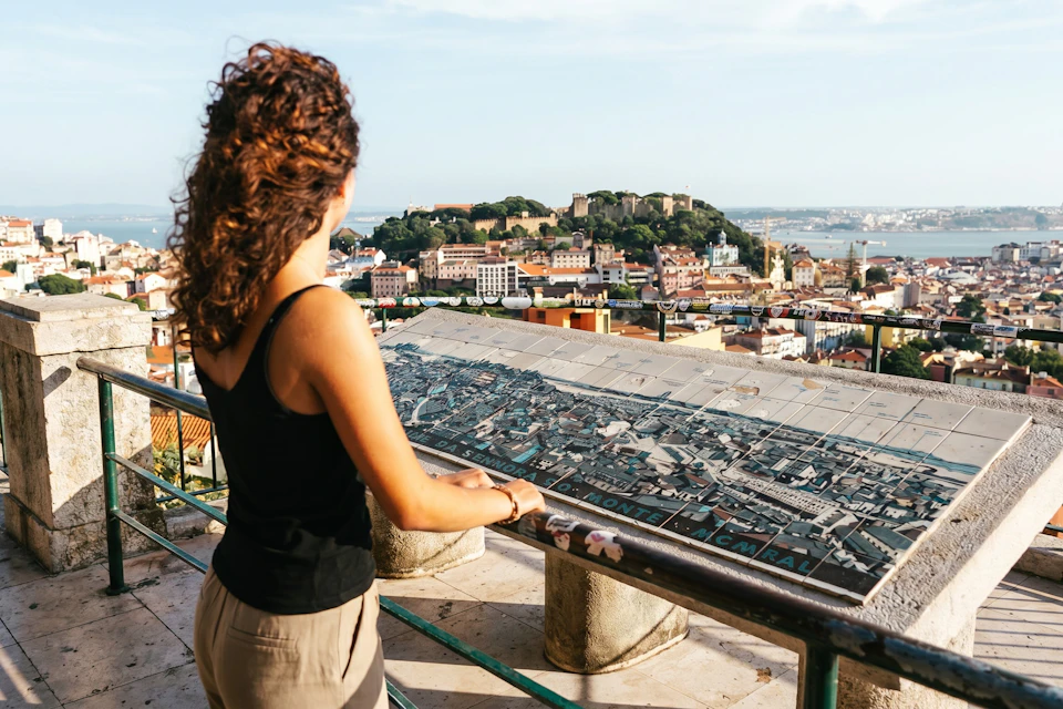 A person with curly hair stands at a railing overlooking a scenic cityscape and a detailed map display in sunny weather.