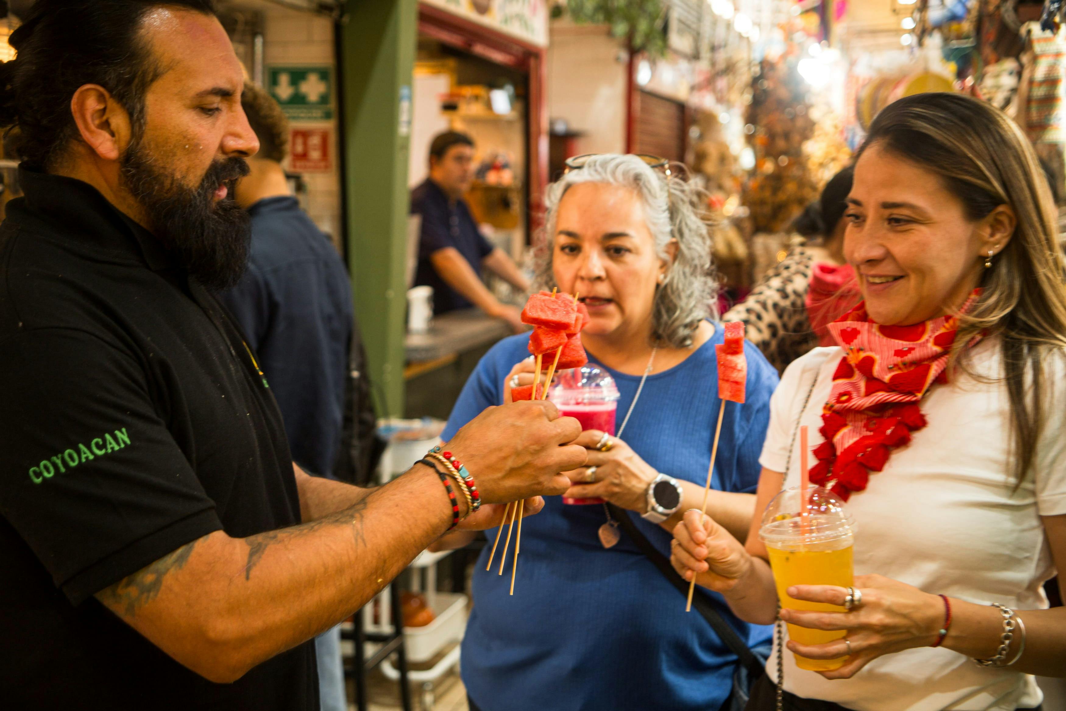 Tres personas comparten en un mercado una brocheta de sandía mientras sostienen bebidas, con puestos de comida visibles al fondo.