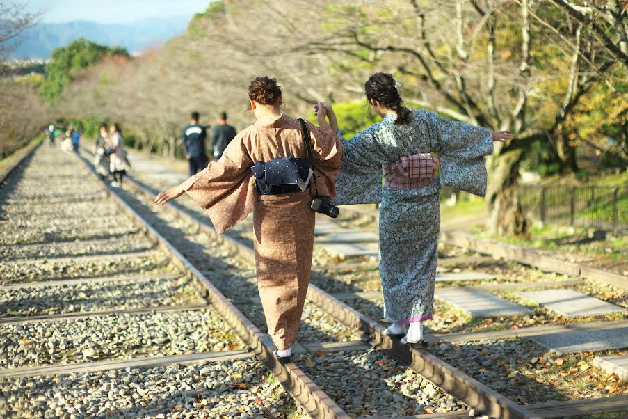 Two individuals wearing traditional Japanese kimonos walking on railway tracks in a scenic area with trees and distant figures.