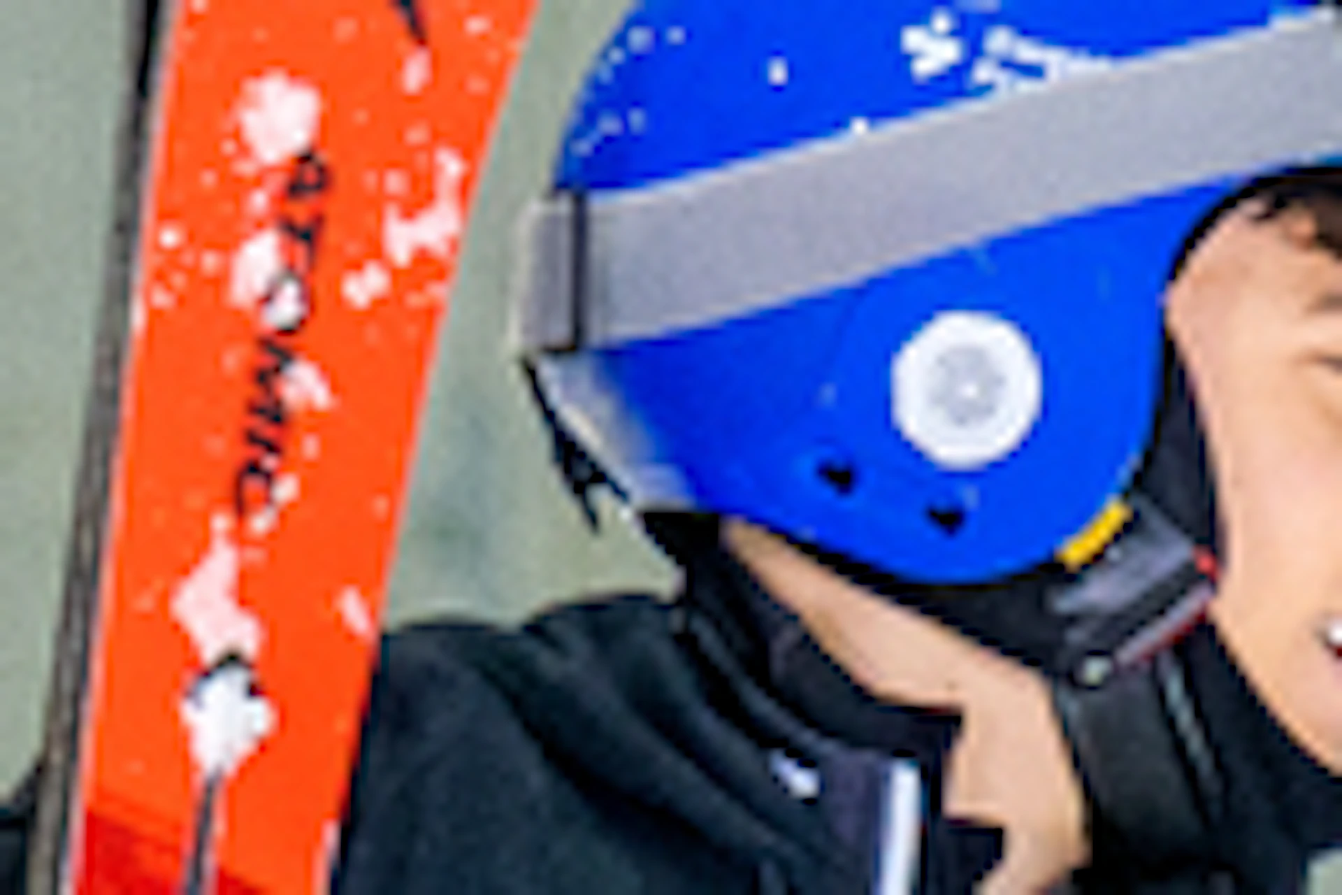 Three people in ski gear walking indoors on artificial snow, carrying skis and poles. Red safety netting in the background.
