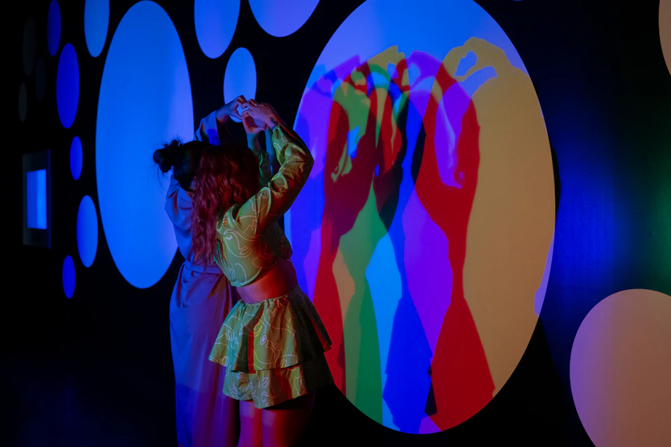 Two people make shadow shapes on a colorful, multi-colored wall display with overlapping blue, green, red, and purple light.