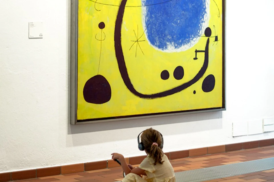 A child in a yellow dress is sitting on the floor, wearing headphones and looking up at a large abstract painting with a yellow background and blue and black shapes.