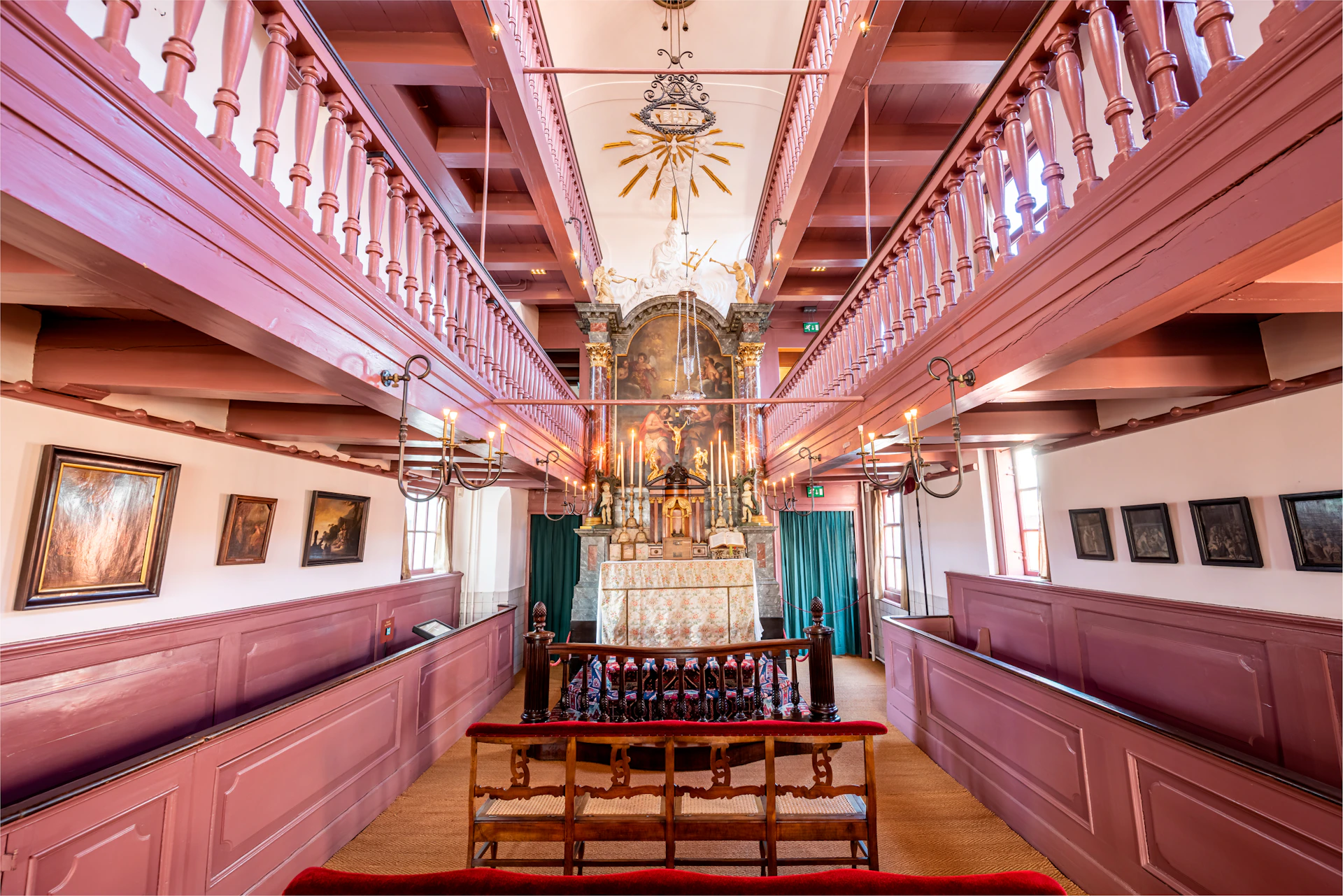 A richly decorated chapel interior with a central altar, ornate chandelier, two levels of pink balustrades, and rows of benches.