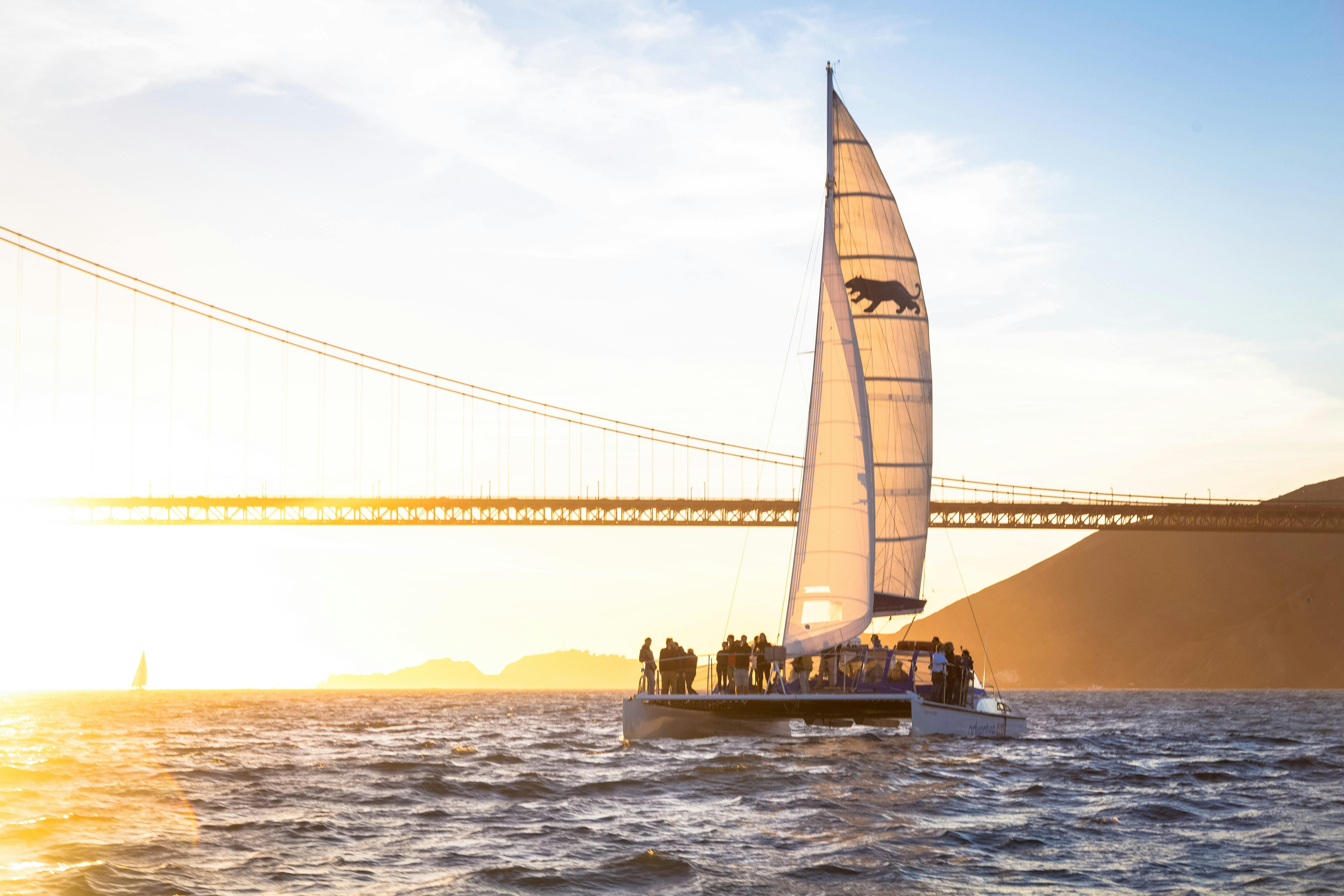 Un catamarano con persone a bordo naviga vicino a un ponte al tramonto. Il sole sta tramontando sull'acqua, proiettando un caldo bagliore.