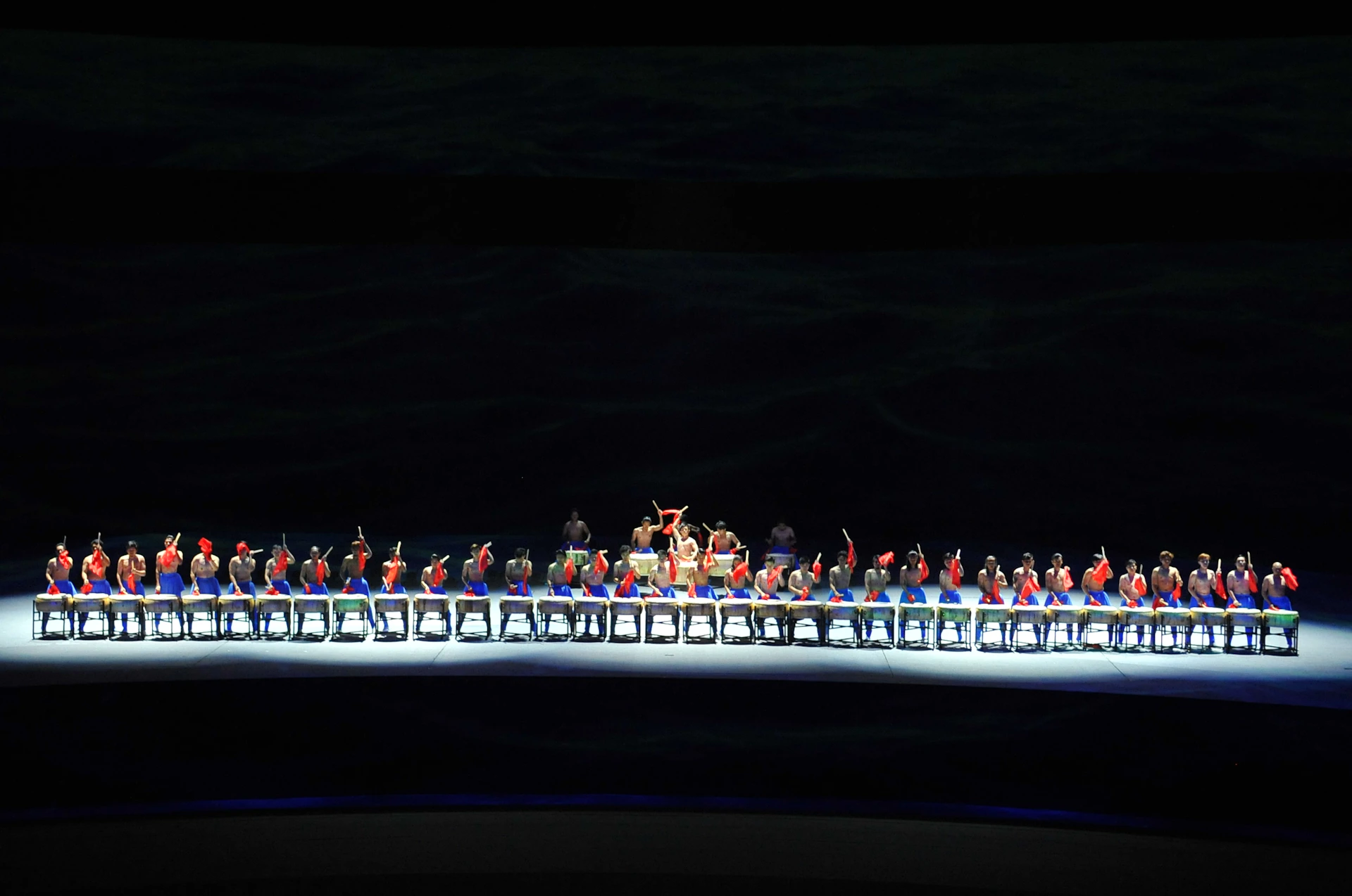 Performers in vibrant outfits on a brightly lit stage, holding props, with a dark background suggesting a theatrical setting.