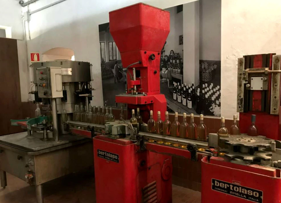 A red Bertolaso bottling machine filling bottles on a conveyor belt in a factory setting. Background shows stacked wine bottles.