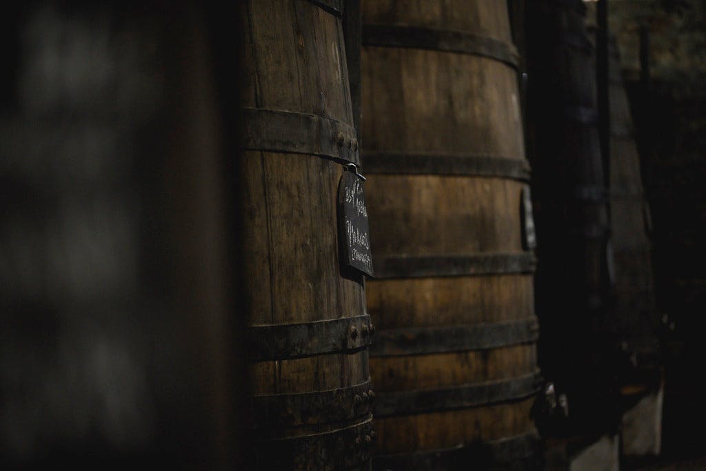 Grote houten vaten met zwarte metalen hoepels, waarvan één met een klein uithangbord, in een schemerige omgeving.