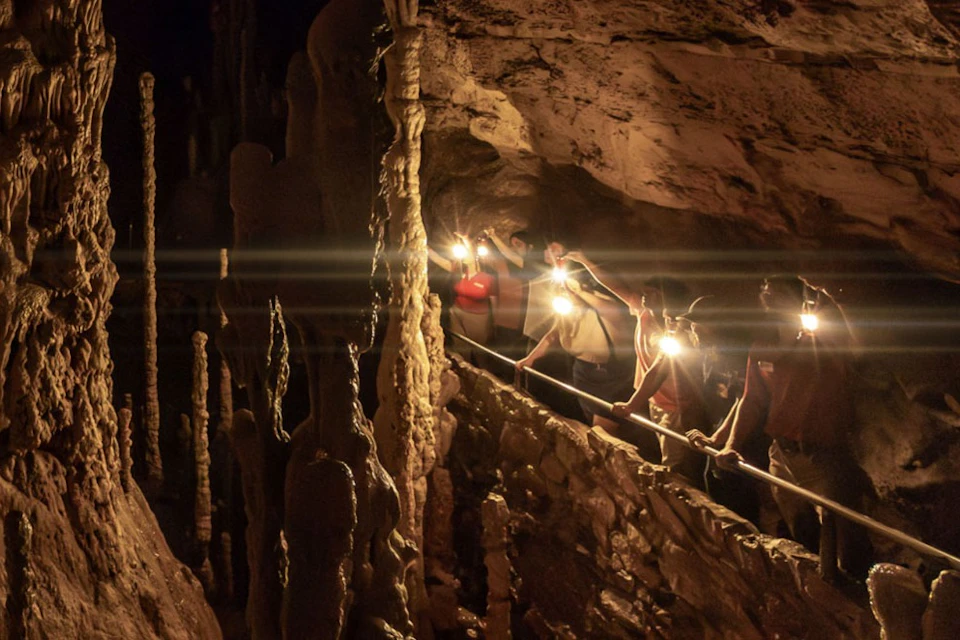 People exploring a cave with stalactites and stalagmites, using hand-held lights along a guided path.