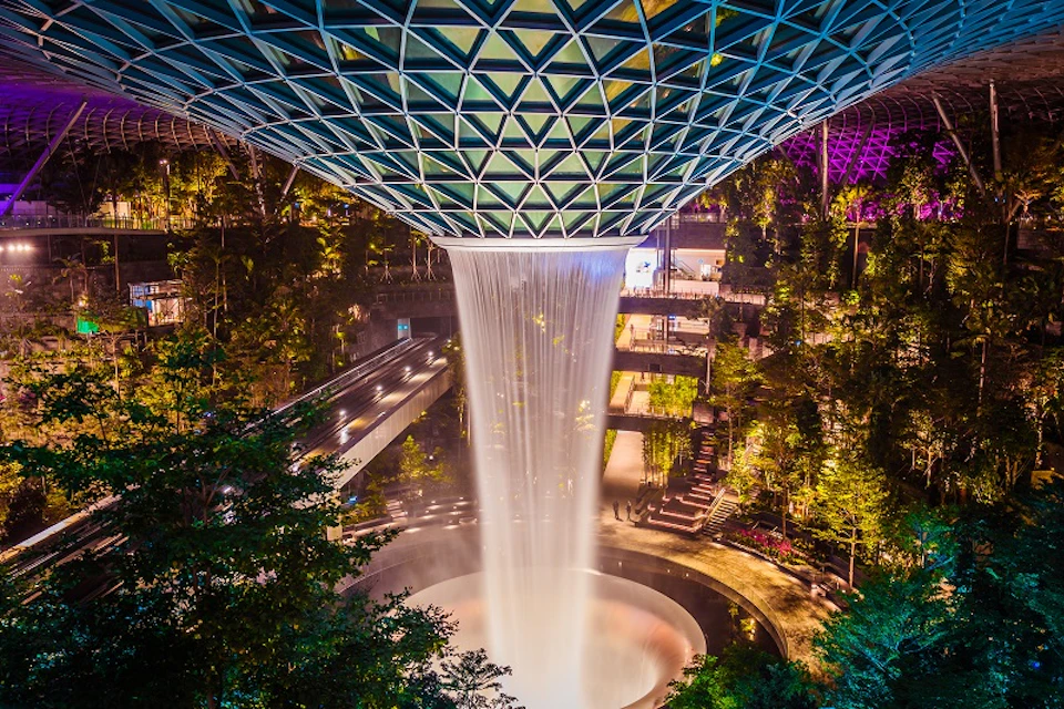 Indoor waterfall with water cascading from a glass dome roof into a circular pool, surrounded by lush greenery and elevated walkways.