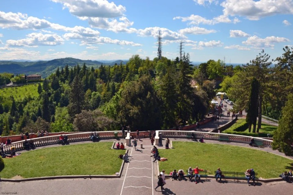 People are sitting and walking in a park area with greenery, pathways, and a scenic view of hills and trees under a partly cloudy sky.