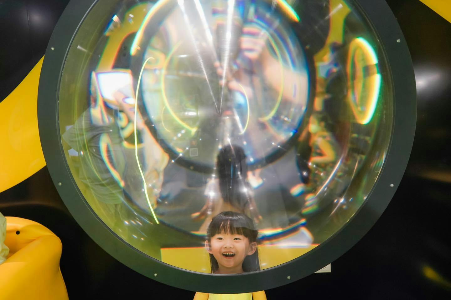 A child smiles behind a large circular glass lens with colorful reflections and distortions.