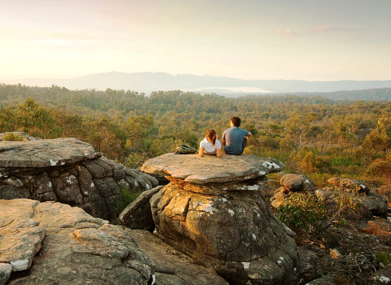 Two people sitting on a large rock, overlooking a vast green forest with distant mountains under a soft, golden sky.