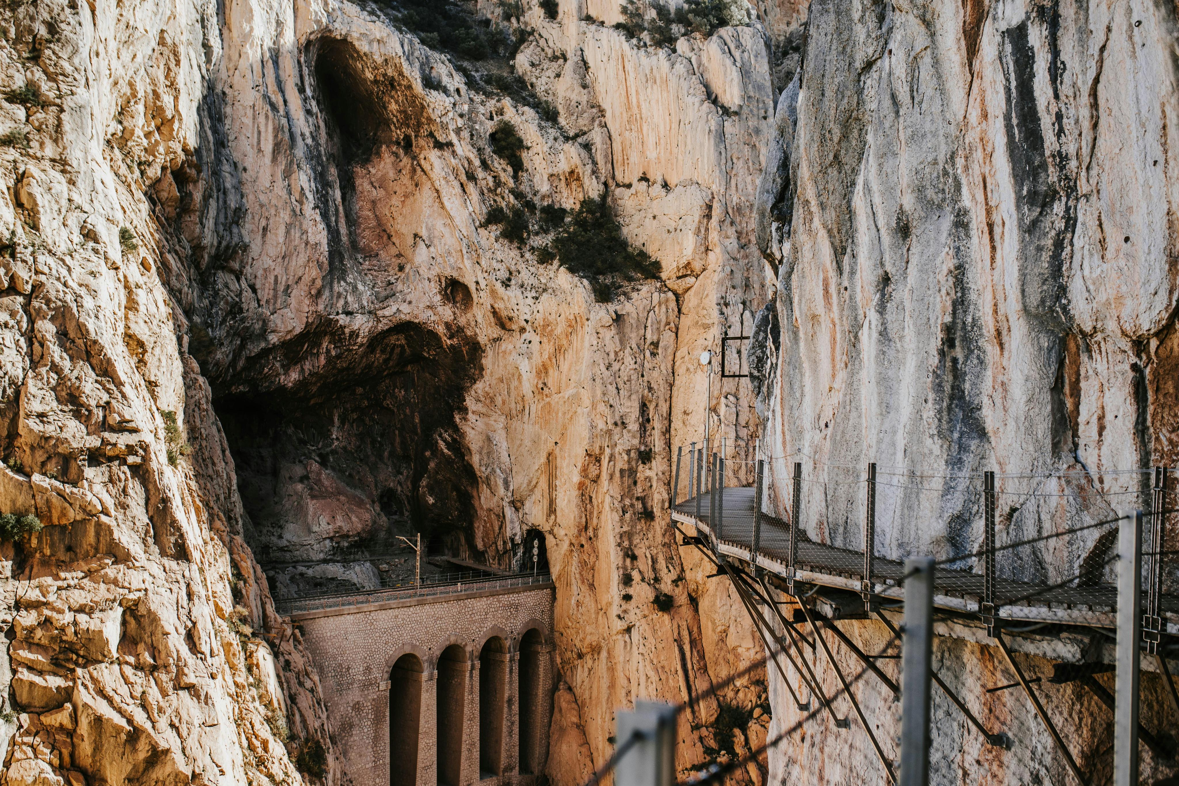 一条狭窄的人行道紧贴着陡峭的岩石悬崖，下面可以看到一座拱形桥。