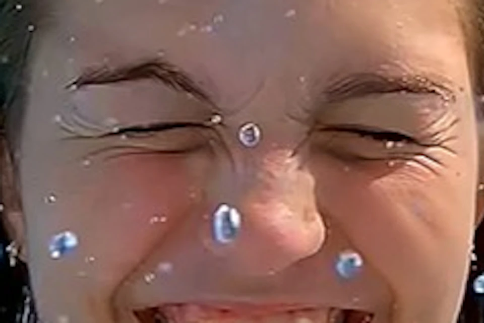 A woman in a blue bikini smiles and squints her eyes, enjoying a water slide ride under a clear blue sky.
