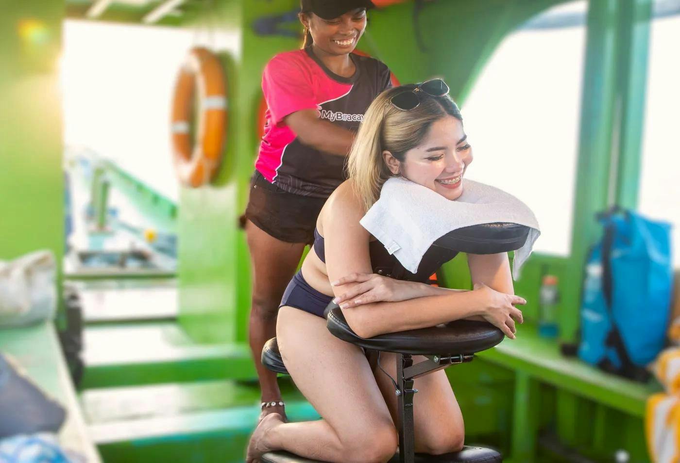 A woman receiving a seated massage from another woman on a boat with green interior.