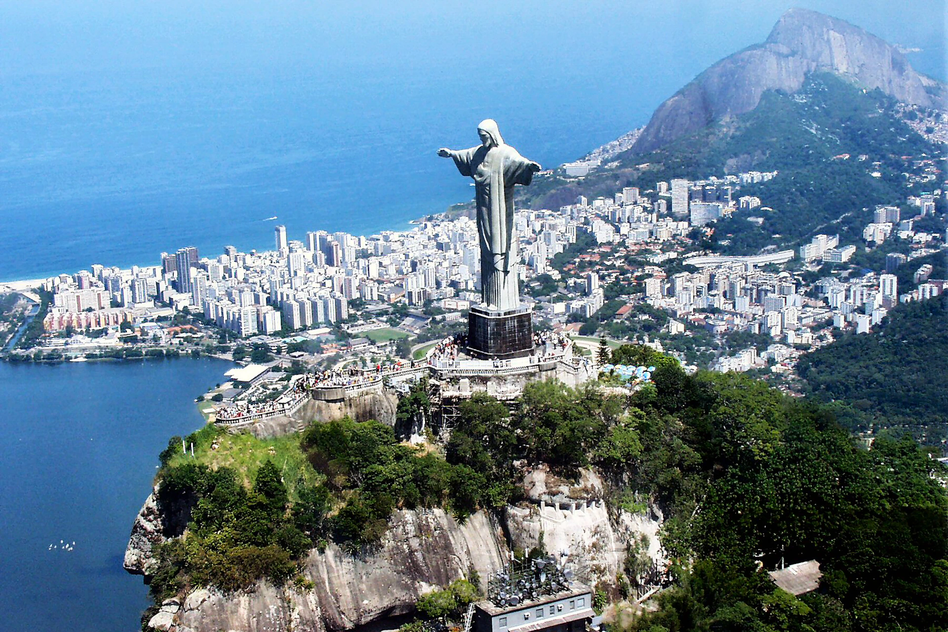 Aerial view of Christ the Redeemer statue on a large hill overlooking a cityscape with numerous buildings and a coastline.