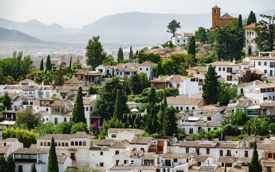 A hillside town with white buildings, tile roofs, and lush greenery. A distant mountain range is visible under a clear sky.