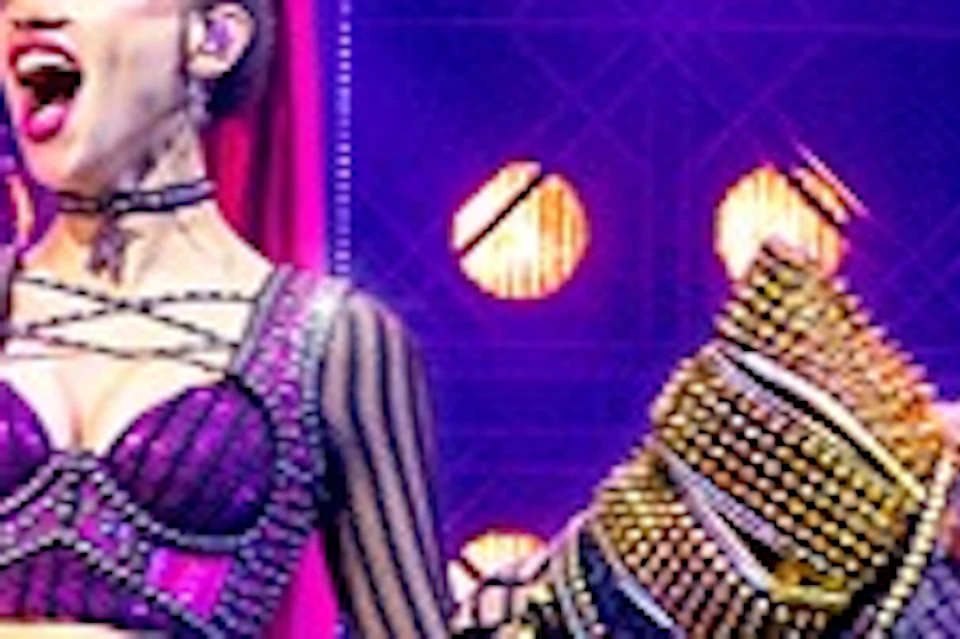 Performers on stage in vibrant costumes singing under purple and yellow lighting, with a lively backdrop and set.
