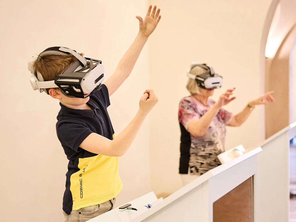 A child and an older adult wearing VR headsets interact in a light room, each gesturing with their hands.