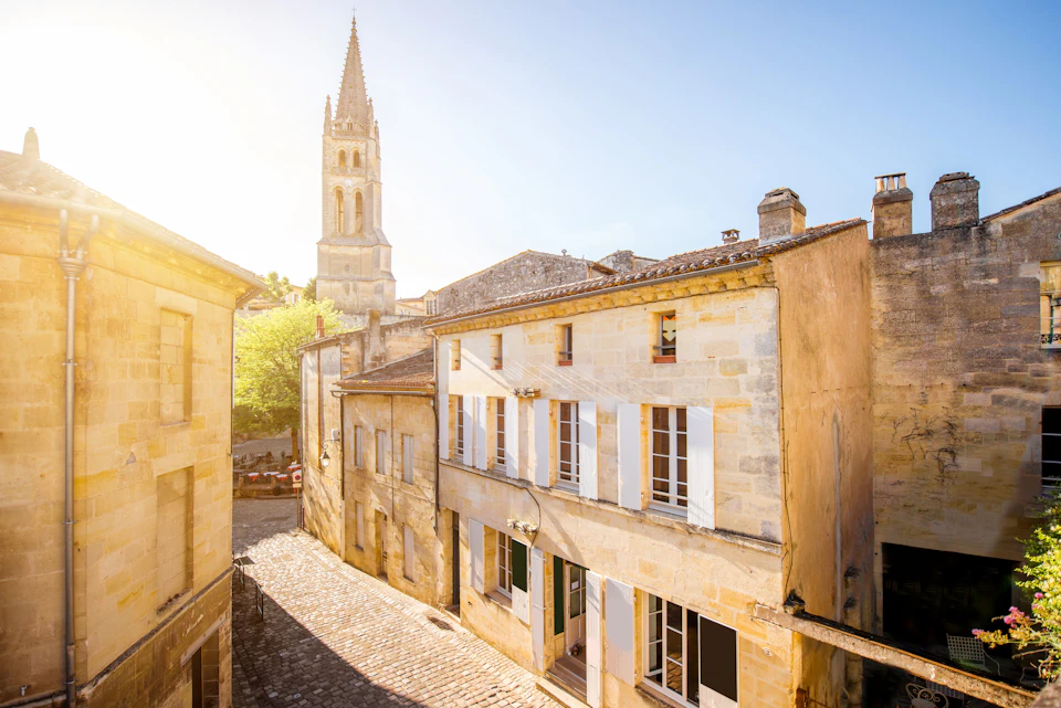 A sunny street scene featuring old stone buildings, white shutters, a cobbled road and a church tower in the background.