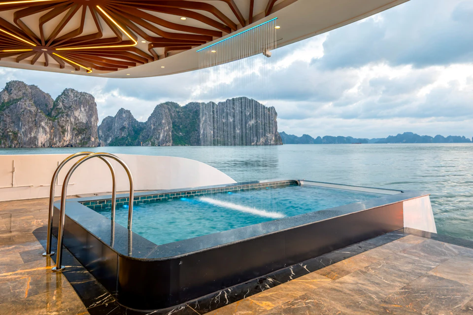 Outdoor rectangular pool with a waterfall feature overlooking a calm bay with limestone islands and a cloudy sky.