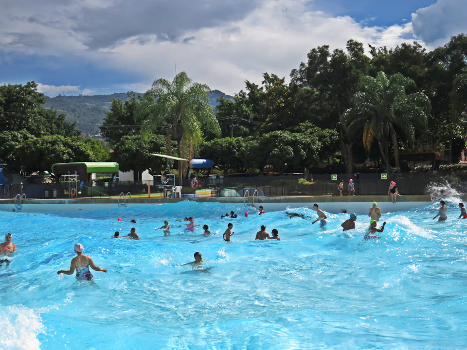 People enjoying a large outdoor swimming pool with gentle waves, surrounded by trees and distant mountains under a cloudy sky.