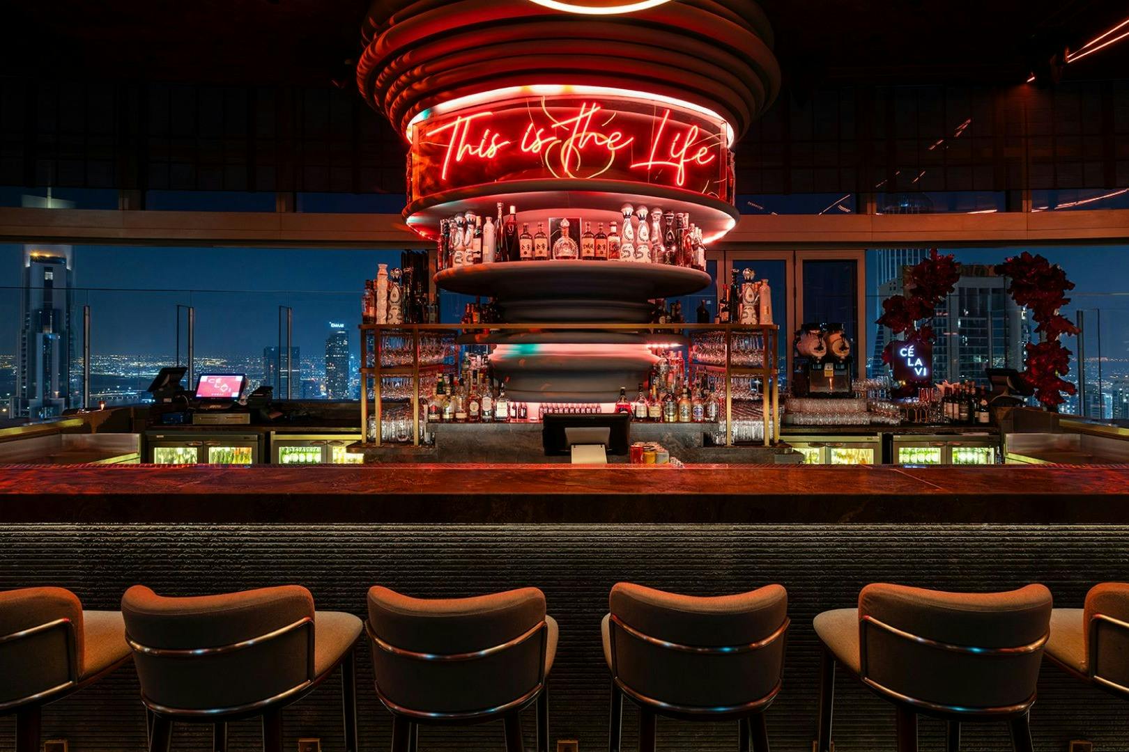 Modern bar with four stools, shelves stocked with bottles, and a neon sign reading "This is the Life" against a cityscape backdrop.