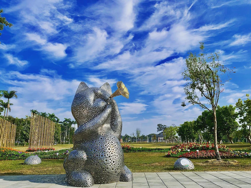 A metal sculpture of a cat playing a trumpet in a park under a blue sky with scattered clouds and surrounding greenery.