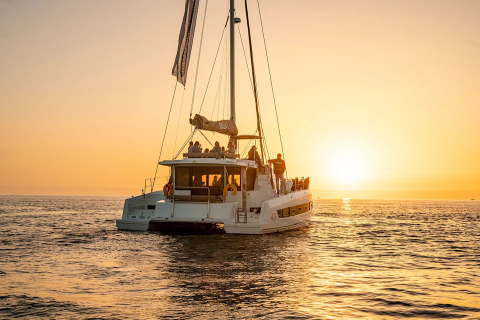 A white sailboat with people aboard sails on calm waters during a vibrant sunset.