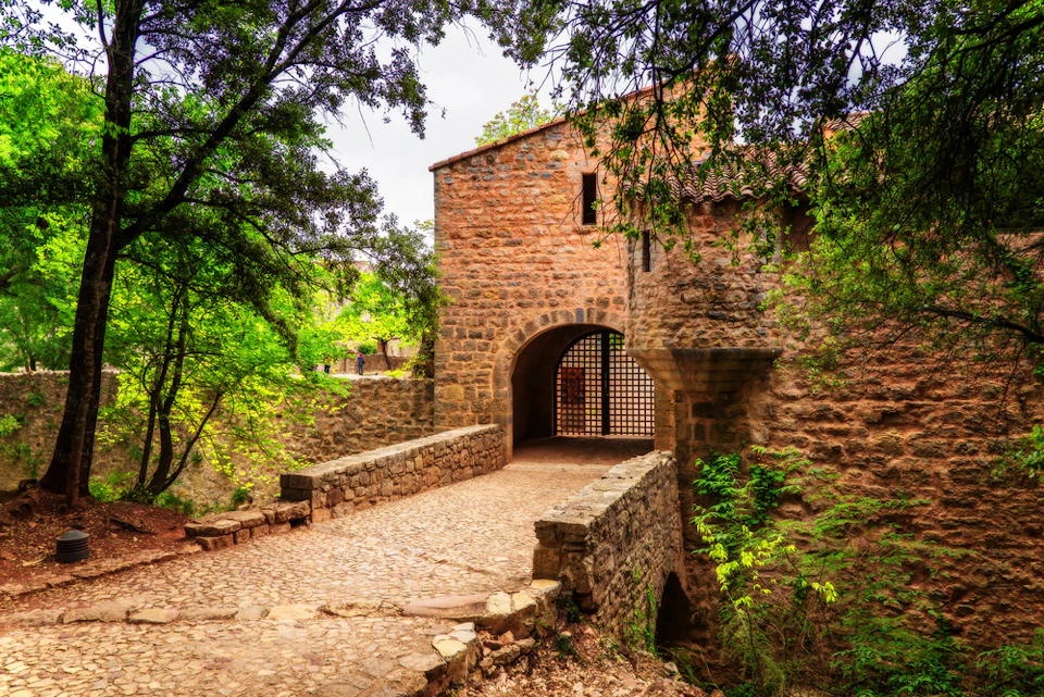 Stone bridge leading to a rustic, arched gateway surrounded by lush green trees and foliage.