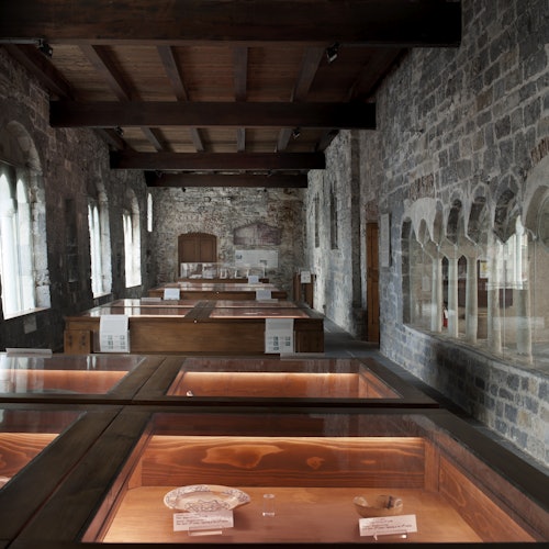 A stone-walled room with wooden display cases showcasing artifacts, featuring arched windows and a wooden ceiling.