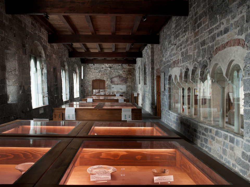 A stone-walled room with wooden display cases showcasing artifacts, featuring arched windows and a wooden ceiling.