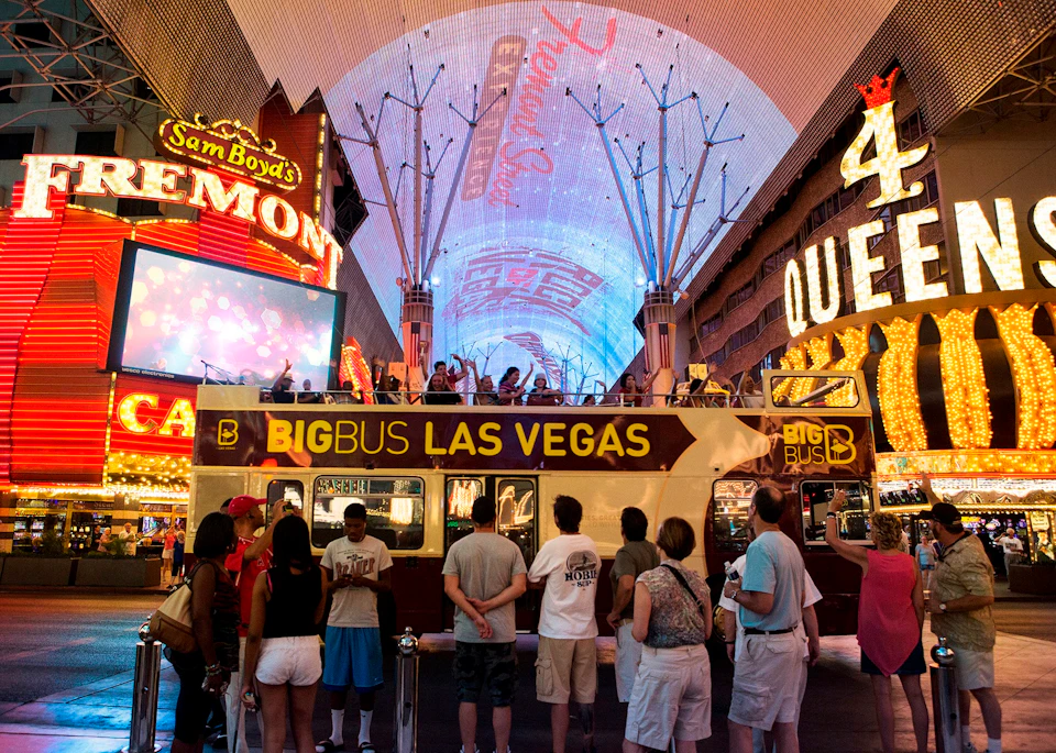 People gather around a "Big Bus Las Vegas" tour bus under illuminated signage at Fremont Street, Las Vegas, at night.