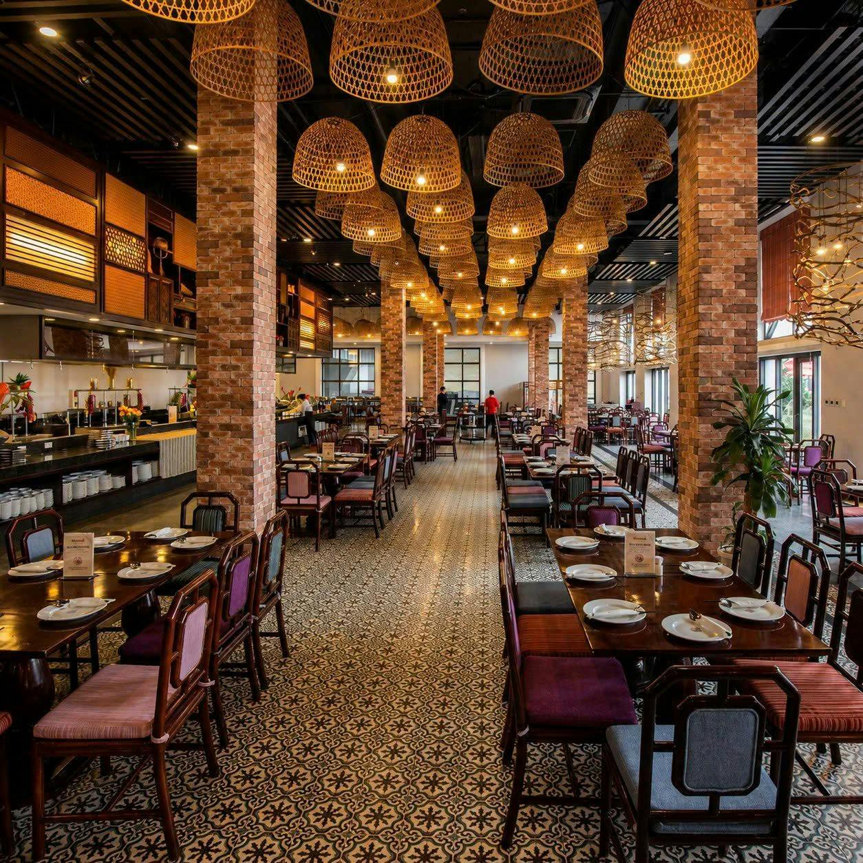 A spacious restaurant with brick columns, patterned tiled floor, and wicker pendant lights. Tables are set with plates.