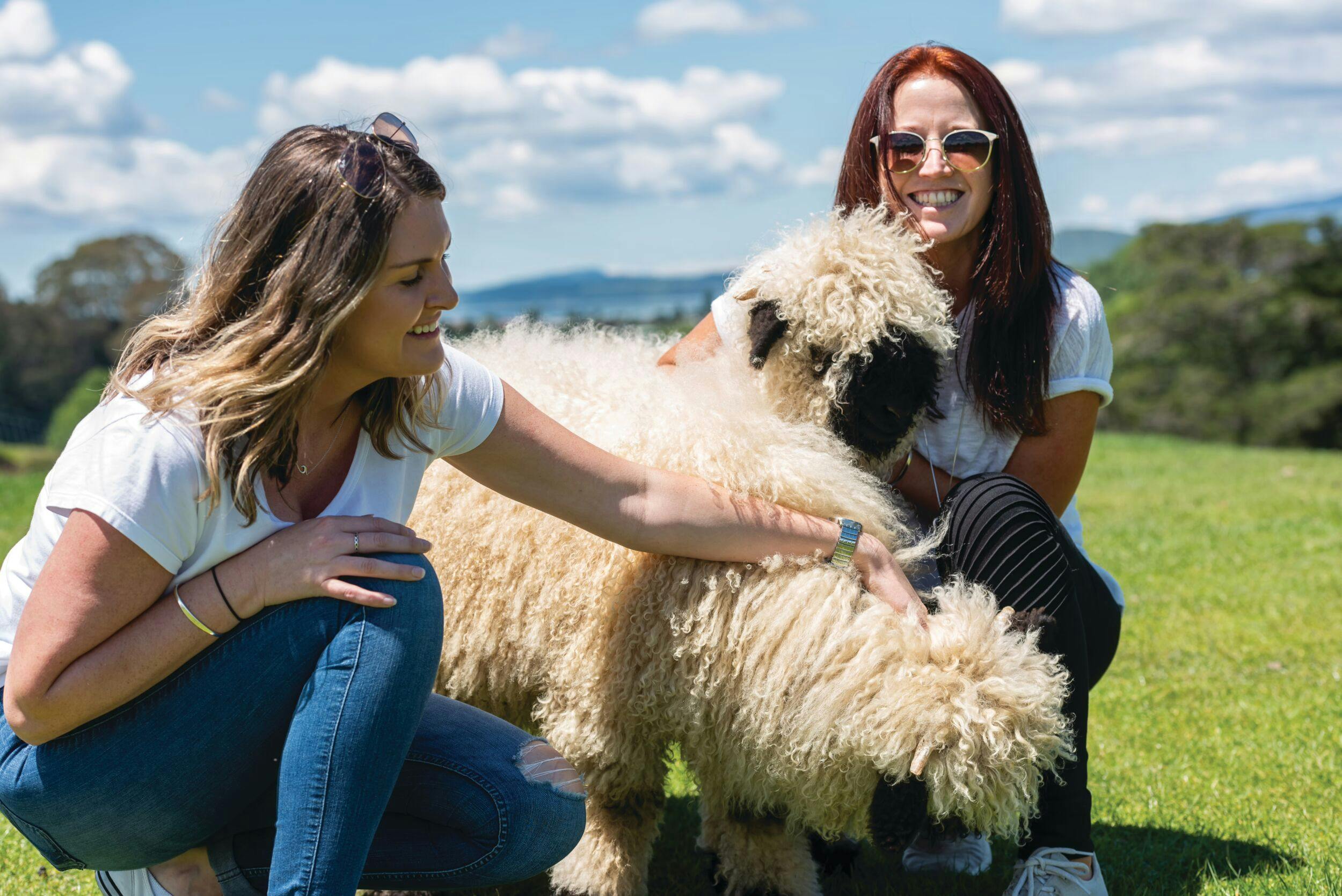 Two women smiling outdoors while petting a fluffy sheepdog in a grassy area under a blue sky with clouds.